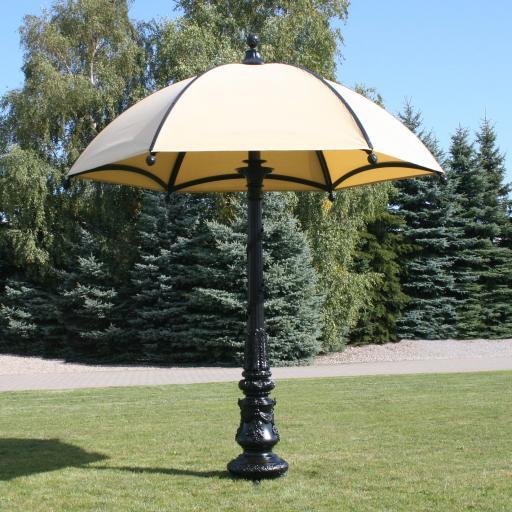 Kousek krajinářské architektury, velký elegantní slunečník v béžovém odstínu na černé ozdobné tyči KM. Deštník je umístěn na volném prostranství, uprostřed přírody.