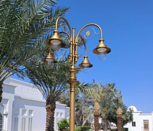 Bohatě zdobená pouliční lampa ve zlatém odstínu se čtyřmi svítidly 04/Aries/A na pozadí palem a bílých budov za slunečného dne.